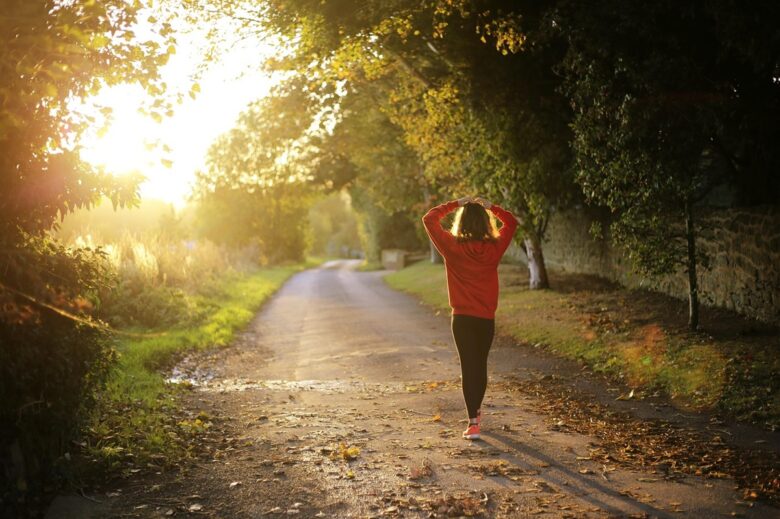更年期の夏を乗り切るため、朝の光の中で木々に囲まれた道をウォーキングする女性。暑熱順化で体を少しずつ動かすセルフケアのイメージ。