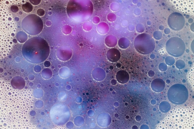 洗剤や柔軟剤に含まれる合成香料の化学成分をイメージした、泡と液体が混ざり合う抽象的なマクロ写真。パープルと白の幻想的な色合い。