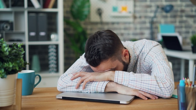 うつむきながら座る男性。肩を落とし、疲労感のある表情で深く考え込んでいる様子。全体的に暗めの色調が、倦怠感やエネルギー不足を強調している。男性更年期障害による身体的な変化を象徴する画像。
