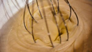 泡立て器で混ぜられている、白っぽい黄色をしたカスタードクリームをクローズアップで捉えたものです。クリームには細かな気泡が含まれており、滑らかな質感が伝わってきます。泡立て器の金属製のワイヤーがクリームの中に深く差し込まれ、動きのある様子が伺えます。これは、ハタダというメーカーの夏季限定スイーツ「おもいでカスタード」のレビュー記事のアイキャッチ画像です。