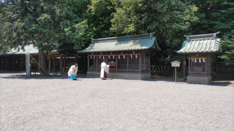 広々とした砂利の境内にある、複数の木造社殿。緑の瓦屋根が特徴的で、手前では白い装束を身につけた二人の人物（神職または巫女）が、社殿に向かって儀式を行っている。周囲は豊かな木々に囲まれ、静かで神聖な雰囲気が漂う神社の様子。