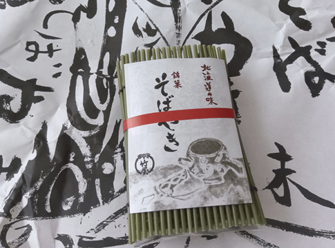 緑色の敷物の上に置かれた、竹屋製菓の銘菓「そばやき」の個包装。黒と白の和紙に包まれ、赤い帯と「北海道の味」の文字が見える。