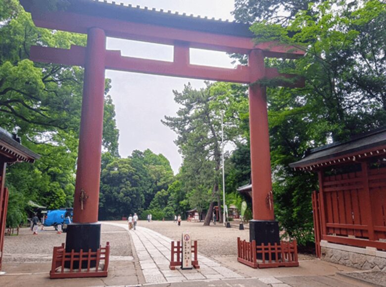 晴れた日に、緑豊かな木々に囲まれた赤色の大きな鳥居が立つ神社の入口。鳥居の下には石畳の参道が奥へと続き、数名の参拝者が歩いている。鳥居の柱元には、黒い柵と赤色の木製の鳥居のような小さな柵が設置されている。
