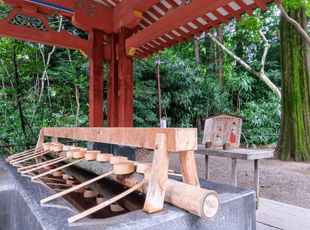 緑豊かな木々に囲まれた神社の手水舎(ちょうずや)。赤色の屋根と柱を持つ建物の中に、石造りの水盤があり、柄杓(ひしゃく)が整然と並べられている。水盤の奥には作法を示すイラスト付きの案内板が置かれており、参拝者が手や口を清める場所であることがわかる。
