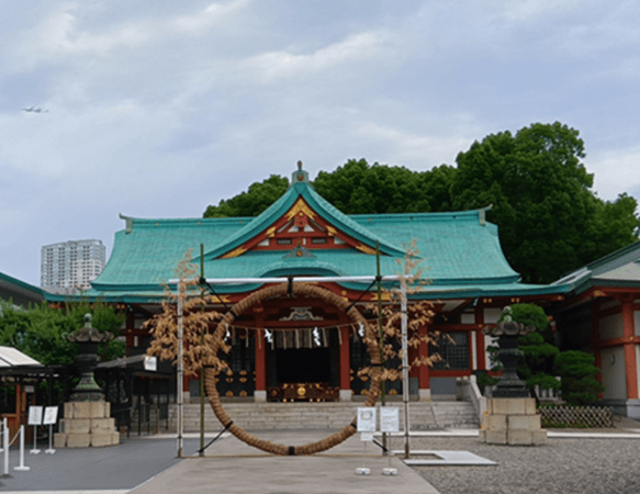 青い空と緑の木々を背景に、神社の拝殿の前に設置された巨大な茅の輪。拝殿は緑色の瓦屋根と朱色の柱が特徴で、茅の輪は太く立派な藁で編まれている。茅の輪の周りには白い紙垂(しで)が飾られ、神聖な雰囲気を醸し出している。右奥にはビルが見え、街中にある神社であることがわかる。