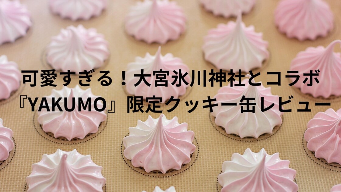 白とピンクのツートンカラーのメレンゲ菓子が、ベージュ色のオーブンシートの上に整然と並んでいるクローズアップ写真。各メレンゲは、下部の平らな部分が白、上部の尖った部分がピンク色で、絞り出しの跡が渦巻き状に残っている。背景はわずかにボケており、同様のメレンゲが多数並んでいる様子が伺える。画面中央には黒い文字で『可愛すぎる！大宮氷川神社とコラボ『YAKUMO』限定クッキー缶レビュー」というブログタイトルが重ねて表示されている。