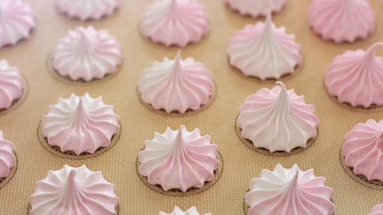白とピンクのツートンカラーのメレンゲ菓子が、ベージュ色のオーブンシートの上に整然と並んでいるクローズアップ写真。各メレンゲは、下部の平らな部分が白、上部の尖った部分がピンク色で、絞り出しの跡が渦巻き状になっている。