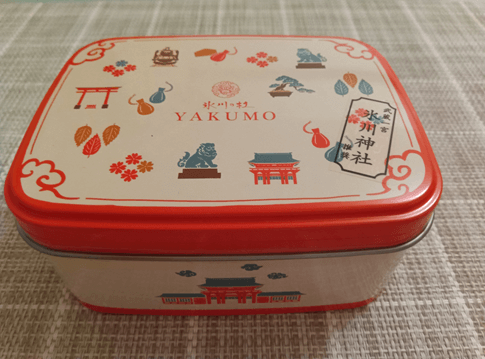武蔵一宮氷川神社と『氷川の杜 yakumo』がコラボした限定クッキー缶の外観。白地に朱色と橙色で鳥居、勾玉、狛犬、盆栽、電車などのイラストが散りばめられ、中央には『氷川の杜 YAKUMO』のロゴと『氷川神社』の札が描かれている。縁と底面は鮮やかな朱色で、全体的に可愛らしく和モダンなデザイン。