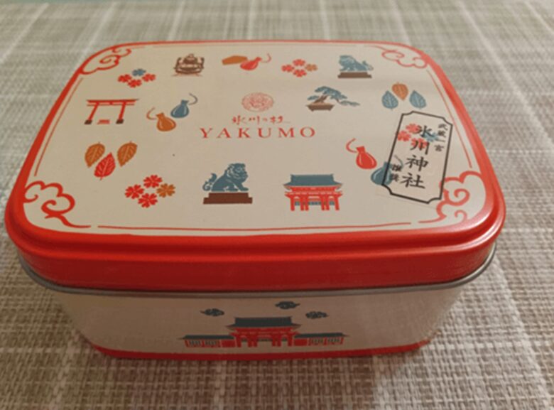武蔵一宮氷川神社と『氷川の杜 yakumo』がコラボした限定クッキー缶の外観。白地に朱色と橙色で鳥居、勾玉、狛犬、盆栽、電車などのイラストが散りばめられ、中央には『氷川の杜 YAKUMO』のロゴと『氷川神社』の札が描かれている。縁と底面は鮮やかな朱色で、全体的に可愛らしく和モダンなデザイン。