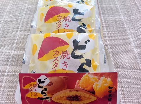 ハタダの秋限定どら焼き「どら一（いち） 焼き芋カスタード」のパッケージが複数個並んでいる写真。手前には商品の断面と焼き芋が写った説明用のPOPがある。
