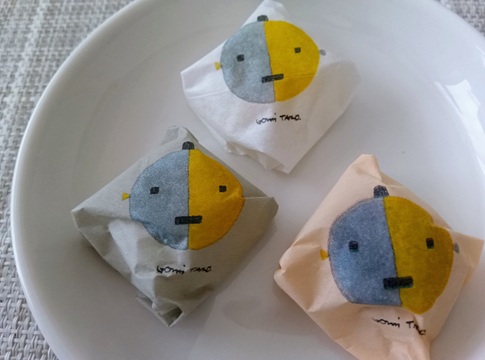 五味太郎のイラストが描かれた廣榮堂きびだんご3種の個包装。白（元祖）、グレー（海塩）、茶色（黒糖）の包み紙が白い皿の上に並んでいる。